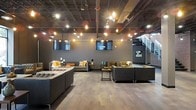 This single-floor space showcases a contemporary industrial aesthetic defined by its exposed ceiling structure, where visible ductwork, support beams, and piping in muted tones create an authentically raw architectural backdrop. Warm Edison bulb string lights hang overhead in a deliberate arrangement, complementing recessed and track lighting to cast an inviting amber glow throughout the room.

The lounge area is furnished with multiple seating arrangements featuring tufted leather sofas and chairs in deep charcoal and brown tones, accented with mustard yellow and orange throw pillows that introduce warmth and contrast. Low-profile industrial-style coffee tables anchor each seating cluster, promoting a social, conversation-friendly layout.

Two wall-mounted flat-screen televisions are positioned prominently along the dark gray accent wall, making the space well-suited for presentations, screenings, or casual viewing. Wide-plank hardwood-style flooring in a warm gray-brown tone grounds the space and adds texture underfoot. A staircase visible in the background hints at additional access points. The overall ambiance is moody, sophisticated, and versatile.