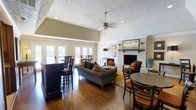 This single-floor space is thoughtfully designed to accommodate both relaxed lounging and casual dining. The room features rich hardwood flooring throughout, which anchors the warm, inviting ambiance. A vaulted ceiling with a textured finish adds a sense of vertical openness, while a centrally mounted ceiling fan ensures comfortable airflow. Recessed lighting complements the natural light that streams in through a series of French doors along one wall, offering views of and access to an outdoor patio or terrace area.

The focal point of the main seating area is a white-painted brick fireplace, topped with a substantial wood mantel and flanked by wall sconces, creating a classic and cozy atmosphere. A gray upholstered sofa, accented with warm-toned throw pillows, faces the fireplace and sits atop a patterned area rug. Additional seating includes accent chairs positioned near the hearth.

Toward the perimeter, bar-height seating and standard dining tables with wooden chairs provide flexible gathering options. A console table against one wall adds functional storage. The overall aesthetic blends traditional architectural details with comfortable, casual furnishings.