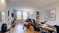 This single-floor residence offers a thoughtfully designed layout that maximizes functionality and comfort. The open-concept living and dining area serves as the heart of the home, featuring warm hardwood flooring, neutral walls, and multiple windows dressed in sheer white curtains that invite generous natural light. The living space is anchored by a navy blue sofa, a glass-top coffee table, and a distinctive gold abstract wall sculpture, while a sleek open shelving unit with wood and metal framing provides both storage and display space. The adjoining dining area accommodates a round wooden table with navy upholstered chairs, ideal for everyday meals or entertaining.

The kitchen is efficiently designed with dark espresso cabinetry, granite countertops, and a full suite of stainless steel appliances including a refrigerator, electric range, and built-in microwave. The hardwood flooring continues seamlessly from the main living area into the kitchen, reinforcing a cohesive aesthetic.

The bedroom is a calm retreat, featuring plush carpeting, large windows with layered blinds and curtains for privacy, and warm ambient lighting. A wooden headboard and coordinating furnishings complete the space.

The bathroom features tile flooring, a dark vanity with a white countertop, a large mirror, and a shower-tub combination, offering a clean and practical design.