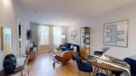 This single-floor residence offers a thoughtfully designed layout that maximizes functionality and comfort. The open-concept living and dining area serves as the heart of the home, featuring warm hardwood flooring, neutral walls, and multiple windows dressed in sheer white curtains that invite generous natural light. The living space is anchored by a navy blue sofa, a glass-top coffee table, and a distinctive gold abstract wall sculpture, while a sleek open shelving unit with wood and metal framing provides both storage and display space. The adjoining dining area accommodates a round wooden table with navy upholstered chairs, ideal for everyday meals or entertaining.

The kitchen is efficiently designed with dark espresso cabinetry, granite countertops, and a full suite of stainless steel appliances including a refrigerator, electric range, and built-in microwave. The hardwood flooring continues seamlessly from the main living area into the kitchen, reinforcing a cohesive aesthetic.

The bedroom is a calm retreat, featuring plush carpeting, large windows with layered blinds and curtains for privacy, and warm ambient lighting. A wooden headboard and coordinating furnishings complete the space.

The bathroom features tile flooring, a dark vanity with a white countertop, a large mirror, and a shower-tub combination, offering a clean and practical design.