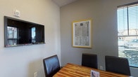 Study Room 1 is a focused, single-level meeting and study space accessed through a glass-paneled door featuring decorative etched signage, providing a semi-private entry that allows visibility into the room. The interior is finished with neutral gray walls and a combination of dark carpet and polished concrete flooring near the entry, giving the space a clean, professional aesthetic.

The central feature of the room is a richly grained, multi-toned wood conference table surrounded by dark leather chairs, offering comfortable seating for small group collaboration or individual study. A wall-mounted flat-screen television is positioned on one wall, making the room well-suited for presentations or media-supported meetings.

Natural light enters through windows fitted with horizontal blinds, offering views of the surrounding exterior while allowing light control. Two framed prints with bold yellow borders serve as wall décor, including a notable reproduction of the Declaration of Independence, adding a distinctive visual element to the space.

A small waste bin is tucked discreetly in the corner, and informational materials are displayed on the table surface. The room's overall layout is efficient and purposeful, designed to support productive meetings, study sessions, or small group gatherings in a tidy, well-appointed environment.