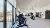 Located at 2700 E Overland Rd in Meridian, Idaho, this generously sized fitness center is designed to deliver a premium workout experience within a bright, modern facility. The single-floor layout is anchored by a dramatic vaulted ceiling that rises to impressive heights, creating an airy, open atmosphere throughout the space. Multiple ceiling fans hang from the peaked roofline, ensuring comfortable air circulation during workouts.

Floor-to-ceiling windows and large glass doors line the walls, flooding the interior with natural light while offering views of an outdoor patio and surrounding community. Roller shades provide adjustable light control without compromising the sense of openness. Light-toned carpet covers most of the floor, while a section with hard flooring near the glass doors adds functional variety.

The equipment selection is extensive and thoughtfully arranged. Cardio options include stationary bikes, treadmills, stair climbers, and rowing machines. A dedicated free weights area features multiple dumbbell racks, barbells, and weight benches, complemented by a power rack and cable machines for strength training. Suspension training equipment and additional multi-use machines round out the offerings.

A geometric accent wall adds a decorative focal point, while built-in shelving provides organized storage. Wall-mounted mirrors in the strength area enhance the sense of space and support proper form during exercise.