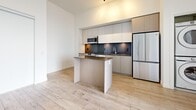 Located at 133 Bronte Rd in Oakville, Ontario, this modern one-bedroom, two-bathroom unit offers a thoughtfully designed open-concept layout with light hardwood flooring and crisp white walls throughout. The spacious living area is bathed in natural light, anchored by floor-to-ceiling windows and a sliding glass door that opens onto a private balcony with breathtaking views of Lake Ontario and the surrounding skyline. The contemporary kitchen is well-appointed with sleek two-tone cabinetry, a dark stone countertop, a central island, and a full suite of stainless steel appliances including a range, microwave, dishwasher, and refrigerator. A dark tile backsplash adds a refined contrast to the light cabinetry. An in-unit stacked washer and dryer is conveniently integrated into the main living area. The generously sized bedroom features a sliding-door closet and tall ceilings, continuing the unit's airy aesthetic. Two bathrooms complete the space — one featuring a glass-enclosed walk-in shower with white subway tiles and a rain showerhead, and a second with wood-toned cabinetry and an accessible grab bar. Four closets provide ample storage throughout the unit, which also includes a dedicated entrance area.