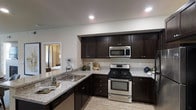 This single-story residence is designed with an open-concept floor plan that promotes seamless flow between the kitchen, dining, and living areas. The entryway hallway is clean and functional, featuring recessed lighting, neutral-toned walls, and classic paneled white interior doors with brushed nickel hardware, providing access to the home's various rooms.

The kitchen is a standout feature of the home, showcasing rich dark espresso-stained cabinetry with ample upper and lower storage. Granite countertops in a light grey and white speckled finish provide generous workspace, complemented by a stainless steel undermount sink with a polished faucet. Stainless steel appliances — including a gas range, over-the-range microwave, and dishwasher — complete the modern kitchen aesthetic. Light hardwood-style flooring runs throughout, adding warmth and continuity across the space.

The open dining area sits adjacent to the kitchen, with a set table visible beneath recessed lighting, and connects naturally to the living room beyond. Large windows in the living area allow natural light to fill the space, creating a bright and welcoming ambiance. Neutral wall tones, recessed lighting, and consistent flooring throughout tie the entire home together in a cohesive, contemporary style.
