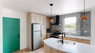 Located at 156 E 900 S in Salt Lake City, Utah, this thoughtfully designed single-floor space offers a contemporary aesthetic with carefully chosen finishes throughout. The kitchen serves as the centerpiece of the unit, showcasing light wood cabinetry with rounded hardware, full-height pantry storage, and stainless steel appliances including a refrigerator and electric range with a sleek black wall-mounted hood. A bold, multicolored terrazzo accent wall adds striking visual texture and frames a window that draws in natural light and views of the surrounding cityscape. The curved kitchen island features a crisp white quartz countertop, an undermount sink with a matte black faucet, and warm wood base cabinetry, all anchored by light hardwood flooring that runs throughout the space. Pendant lighting with amber globe fixtures adds a warm, inviting glow. The bathroom continues the design-forward approach with floor-to-ceiling blush ceramic tile, a light wood vanity with a white countertop, an arched mirror, matte black fixtures, and a full soaking tub with shower. A private balcony with black metal railings overlooks a canopy of mature trees, providing a serene outdoor retreat. A vibrant green entry door adds a bold pop of color to the interior.