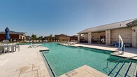 Located in Celina, Texas, this community amenity center offers a resort-style outdoor experience designed for relaxation and recreation. The centerpiece of the space is a generously sized swimming pool with crystal-clear turquoise water, featuring both shallow wading areas and deeper swimming sections, clearly marked for safety. In-pool tanning ledges with built-in jet features provide a luxurious touch, inviting residents to lounge directly in the water. An ADA-compliant pool lift ensures accessibility for all users.

The pool deck is finished with light-toned pavers that surround the water on multiple sides, providing ample space for lounging. Rows of cushioned chaise lounge chairs and patio seating are arranged throughout the deck, with several shaded umbrella stations offering relief from the Texas sun.

The facility is anchored by a single-story brick building with a standing seam metal roof, characteristic of contemporary Texas architecture. A covered outdoor patio with ceiling fans extends from the structure, creating a comfortable shaded gathering area adjacent to the pool. Large windows along the building's exterior allow natural light to flow between the indoor and outdoor spaces.

The entire pool area is enclosed by wrought iron fencing, providing security while preserving open sightlines to the surrounding neighborhood. The space is well-suited for both casual leisure and community gatherings.