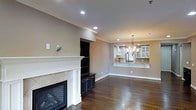 This single-floor residence showcases an open-concept layout that seamlessly connects the living area to the kitchen and dining space. The living room serves as the focal point of the home, anchored by a classic fireplace with a white mantel and tile surround, flanked by a built-in entertainment unit with dark cabinetry, offering both display and storage space. Rich, dark hardwood floors run throughout, adding warmth and a sense of continuity between rooms.

Crown molding lines the ceilings, lending a refined architectural detail to the space. Recessed lighting provides even illumination throughout, complemented by a decorative chandelier that marks the dining area and adds an elegant touch.

A raised breakfast bar with granite countertops creates a natural transition into the kitchen, which features white cabinetry, glass-front upper cabinets, a range hood, and stainless steel appliances. The kitchen's bright, clean aesthetic contrasts pleasantly with the warmer tones of the living space. The neutral wall tones throughout tie the entire open-plan area together, creating a cohesive and inviting atmosphere.