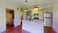 This single-floor unit showcases a thoughtfully designed interior with a contemporary aesthetic carried consistently throughout the space. Warm-toned hardwood flooring runs throughout, anchoring each area with a cohesive, inviting feel.

The kitchen serves as the centerpiece of the living area, designed in an open-concept layout that promotes connectivity and functionality. A large central island with a light quartz countertop and an undermount sink with a matte black faucet provides both workspace and casual seating. Pendant lighting with mid-century modern styling hangs above the island, complemented by a multi-directional track light fixture overhead. Flat-panel grey cabinetry extends from floor to ceiling, offering generous storage, while a herringbone-patterned tile backsplash adds visual texture. Stainless steel appliances, including a range, microwave, and refrigerator, complete the kitchen.

The bathroom includes a full tub-shower combination, a vanity with a stone-topped surface, and coordinating matte black hardware. A stacked washer and dryer unit is conveniently integrated into the bathroom, adding practical in-unit laundry access.

A bedroom features a large window with urban views, allowing natural light to fill the space.