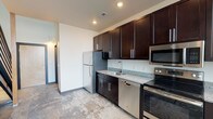 This two-story, 1-bedroom, 1.5-bathroom loft offers a thoughtfully designed living space that blends modern finishes with an open, airy layout. The kitchen is well-appointed with rich dark espresso cabinetry, granite countertops, stainless steel appliances including a refrigerator, dishwasher, microwave, and electric range, plus a breakfast bar with a built-in wine rack — ideal for casual dining and entertaining. Pendant lighting adds a stylish industrial touch above the bar area. The main living area features warm hardwood flooring and soaring vertical wall space, giving the home an expansive, loft-like feel. A staircase with metal railings leads to the upper-level loft, which serves as the bedroom and overlooks the main floor. A skylight on the upper level brings in natural light. The full bathroom is located on the main level and includes hardwood flooring, a tub/shower combination, a granite-topped vanity, and dark cabinetry consistent with the rest of the home. A convenient half-bathroom is also available, finished with a granite vanity countertop and a large mirror. Throughout the unit, cohesive design choices — including granite surfaces and dark cabinetry — create a polished, modern aesthetic.