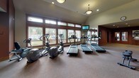 This single-level fitness center is thoughtfully designed to accommodate a wide range of workout needs within a spacious, open layout. The main cardio area is anchored by a row of treadmills and elliptical machines positioned along a generous bank of windows, allowing abundant natural light and outdoor views to create an energizing atmosphere. Vaulted ceilings with pendant lighting and recessed fixtures add an open, airy quality to this zone, while a wall-mounted television provides entertainment during workouts.

The strength training area features a variety of cable and weight machines, along with a flat bench for free weight exercises. Deep burgundy accent walls contrast with neutral carpeting throughout, lending the space a polished yet functional character. Strategic placement of wall mirrors enhances the sense of depth and allows users to monitor their form.

A dedicated functional training nook houses stability balls on a vertical rack, medicine balls, a BOSU balance trainer, and a full dumbbell rack with weights ranging across multiple sizes. Warm wood-trimmed double doors connect this area to an adjacent common space, reinforcing an integrated, resort-style feel.

A corridor off the main floor provides access to a wall-mounted water fountain and additional equipment, completing a comprehensive and well-organized fitness facility.