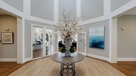 This single-story clubhouse presents a thoughtfully designed communal space that balances elegance with functionality. The grand foyer immediately establishes the tone, showcasing soaring ceilings, warm hardwood-style flooring, and a striking round accent table topped with an oversized floral arrangement. A large-scale ocean-inspired artwork adorns the wall, complementing the soft gray color palette carried throughout the space.

The main lounge area offers generous seating with neutral sofas accented by rich emerald green and patterned throw pillows. A classic white fireplace surround with a wall-mounted television serves as a focal point, while abundant natural light streams through large windows offering outdoor views. Nearby, a secondary seating arrangement with navy accent chairs provides a more intimate gathering spot.

The business center features an L-shaped granite countertop workstation accommodating multiple computer stations, complemented by botanical artwork in gold frames. The game room includes a full-size billiards table, wall-mounted television, and high-top seating alongside ample natural light from a bank of windows.

The kitchen is finished with white shaker cabinetry, dark granite countertops, a gray subway tile backsplash, stainless steel appliances, and a sink-equipped island, making it well-suited for community events and gatherings.