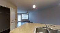 This single-floor residence offers a well-designed open-concept layout that seamlessly connects the kitchen and living areas. The kitchen features granite-style countertops, a stainless steel sink, and a pendant light that adds a contemporary touch overhead. The countertop extends as a functional peninsula, creating a natural division between the kitchen and the main living space while maintaining an open, airy feel.

The living area is finished with dark hardwood-style flooring that provides a rich contrast against the cool, light gray walls. A ceiling fan is centrally positioned, ensuring comfortable airflow throughout the space. Natural light pours in generously through a large window and a glass-paneled door that opens to what appears to be an outdoor balcony or patio, bringing in views of surrounding greenery. The combination of natural light and neutral tones gives the space a calm, inviting ambiance.

The layout is practical and efficient, making the most of the available space while maintaining a clean, uncluttered aesthetic. Overall, this single-level residence balances modern finishes with functional design, making it a comfortable and well-lit living environment.