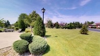 This single-floor property is distinguished by its exceptionally well-maintained outdoor grounds, which form a striking first impression. The expansive lawn is carefully manicured, presenting a rich, even green carpet that stretches generously across the property. Neatly trimmed shrubs and rounded hedges line the borders, contributing a sense of structure and intentional landscaping design. Tall evergreen trees and a variety of deciduous specimens are thoughtfully placed throughout the grounds, providing shade, visual depth, and year-round greenery. A classic black lamp post stands prominently along a gravel-edged planting bed, adding a timeless, decorative element that enhances the overall curb appeal. In the background, a brick-toned building is partially visible through the tree line, suggesting a well-established and cohesive property setting. The open sky above, with its light cloud cover, floods the outdoor space with natural light, lending an airy and inviting atmosphere. The overall ambiance of the grounds is one of careful upkeep and classic charm, making this property an appealing and polished space suited for those who appreciate well-designed, serene outdoor environments.