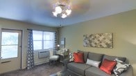 This single-floor space offers a comfortable and thoughtfully arranged living area that combines functionality with decorative charm. The living room serves as the focal point, featuring a multi-bulb ceiling fan light fixture that provides warm, ambient illumination. The walls are finished in soft, neutral tones that create a calm and welcoming atmosphere throughout. A plush gray sofa anchors the seating area and is accessorized with vibrant red and silver accent pillows, adding a pop of color and visual interest. A patterned area rug grounds the space and complements the overall color palette. Decorative wall art with a geometric Moroccan-inspired motif adds a stylish touch to the room. Natural light filters in through a window dressed with bold black-and-white patterned curtains, contributing to the bright and airy feel of the space. A mirrored vanity desk and chair tucked near the window offer a practical workspace or dressing area. A wall-mounted HVAC unit ensures year-round climate comfort. The carpeted flooring adds warmth and softness underfoot, tying together the cozy and livable character of this well-appointed space.