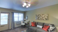 This single-floor space offers a comfortable and thoughtfully arranged living area that combines functionality with decorative charm. The living room serves as the focal point, featuring a multi-bulb ceiling fan light fixture that provides warm, ambient illumination. The walls are finished in soft, neutral tones that create a calm and welcoming atmosphere throughout. A plush gray sofa anchors the seating area and is accessorized with vibrant red and silver accent pillows, adding a pop of color and visual interest. A patterned area rug grounds the space and complements the overall color palette. Decorative wall art with a geometric Moroccan-inspired motif adds a stylish touch to the room. Natural light filters in through a window dressed with bold black-and-white patterned curtains, contributing to the bright and airy feel of the space. A mirrored vanity desk and chair tucked near the window offer a practical workspace or dressing area. A wall-mounted HVAC unit ensures year-round climate comfort. The carpeted flooring adds warmth and softness underfoot, tying together the cozy and livable character of this well-appointed space.