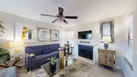 This well-appointed single-floor residence offers a thoughtfully designed layout that balances comfort and style. The living room serves as the centerpiece of the home, featuring light wood-look flooring, a textured ceiling, and a ceiling fan overhead. A classic fireplace with a white mantel anchors one wall, complemented by a wall-mounted television above. Natural light filters through plantation-shuttered windows, illuminating the space and highlighting the curated décor, including framed artwork and gold-accented furnishings that lend a warm, sophisticated character throughout.

The bedroom is a standout space, distinguished by a dramatic deep navy accent wall adorned with framed artwork. Plush carpeting and a ceiling fan contribute to a restful atmosphere, while additional windows continue the theme of natural light.

A sliding glass door leads from the kitchen area to a private outdoor patio, where a small bistro seating area rests on a patch of greenery — ideal for enjoying fresh air. The stainless refrigerator and vertical blinds frame this seamless indoor-outdoor connection.

The bathroom is efficiently designed, featuring a wide vanity with an integrated sink and a stacked washer and dryer unit tucked neatly within, providing the convenience of in-unit laundry. Together, these spaces form a cohesive, comfortable, and stylishly finished home.