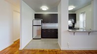 This single-floor unit is defined by its warm, classic parquet wood flooring, which runs consistently throughout the living spaces and bedrooms, adding a timeless character to each room. The layout, as seen in the 3D floor plan, includes a living area, kitchen, multiple bedrooms, and a bathroom, all arranged in a functional configuration.

The kitchen stands out with its contemporary dark cabinetry, granite-style countertops, stainless steel refrigerator, and gas range. Large format tile flooring in the kitchen provides a clean contrast to the parquet found in the rest of the unit. Recessed flush-mount ceiling lights illuminate the space effectively.

The living and bedroom areas feature white walls, flush-mount ceiling lighting, and multiple windows fitted with horizontal blinds, allowing natural light to filter in from multiple directions. Baseboard heating units are present in several rooms, and at least one window accommodates an air conditioning unit. Bedrooms include closets with built-in hanging rods, providing practical storage.

The bathroom is finished in white tile, featuring a full tub and shower combination, chrome fixtures, and a window for natural ventilation. The overall ambiance of the unit is clean, neutral, and move-in ready.