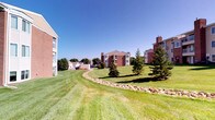 This single-floor residence is part of a classic brick multi-building community set among expansive, well-manicured green lawns accented by mature evergreen trees and decorative stone landscaping borders. The exterior grounds offer a park-like setting with open sightlines and a serene, suburban atmosphere under wide open skies.

Inside, the bathroom is thoughtfully appointed with a full-size vanity featuring a wide basin sink, chrome fixtures, and a large lighted mirror that brightens the space. Neutral tones throughout the bathroom create a clean, understated aesthetic. The tub and shower combination is tiled in classic light pink square tiles with a dual showerhead setup — both a fixed overhead and a handheld — providing versatility and comfort. A curtain rod and shower curtain offer privacy, while chrome towel bars provide convenient storage.

A standout functional feature visible from the bathroom is the stacked washer and dryer unit, housed in a dedicated closet — offering the convenience of in-unit laundry. Dark wood-toned doors and trim provide a warm contrast against the light neutral walls and tile throughout the space. The overall ambiance is practical, clean, and comfortable, making this a functional and well-equipped residence within a landscaped community setting.