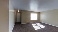 This single-floor residential space presents a straightforward and functional layout designed for comfortable everyday living. The main living area is finished with dark wall-to-wall carpeting that adds warmth underfoot, while the neutral beige walls provide a versatile backdrop suited to a variety of interior styles. A large window allows natural light to filter into the room, casting bright patches across the floor and creating an inviting atmosphere throughout the day. The textured popcorn ceiling adds a classic residential character to the space. A hallway visible from the main room leads further into the interior, suggesting additional rooms and providing a sense of depth and connectivity within the floor plan. The clean white baseboards offer a crisp contrast against the darker carpet and tan walls. The space is presented in a vacant state, allowing the architectural layout and features to be clearly observed. Overall, the unit offers a modest yet practical living environment with a neutral palette that lends itself well to personalization.
