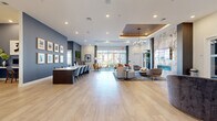 This elegantly designed community amenity space at 100 Forge Circle in Lawrence Township, NJ offers a sophisticated open-plan layout spread across a single expansive floor. Light hardwood flooring flows throughout, anchoring the modern aesthetic and tying together the various functional zones seamlessly. A striking tray ceiling with warm wood inlay and a sculptural branch-style chandelier serves as a dramatic focal point, adding depth and architectural interest overhead. The lounge area is anchored by a floor-to-ceiling marble-surround linear fireplace paired with a mounted flat-screen display, surrounded by plush, contemporary seating including curved accent chairs and a generous sectional sofa atop a patterned area rug. A dedicated conference and collaboration zone features a long dark-wood table flanked by blue velvet chairs, set against a bold slate-blue feature wall adorned with a curated gallery of framed artwork. Oversized floor-to-ceiling windows flood the interior with natural light and offer views of the surrounding community courtyard, creating a bright and welcoming atmosphere. Additional seating vignettes with leather and upholstered chairs are thoughtfully arranged throughout. A separate office or meeting room is accessible via glass-paneled doors, adding versatility to the overall space.
