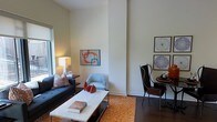 This single-floor space makes a strong first impression with its notably high ceilings and clean, neutral walls that create an open, airy atmosphere. The living area is thoughtfully appointed with a tufted navy sofa accented by patterned throw pillows, anchored by a sleek marble-topped coffee table resting on a vibrant orange area rug. The warm tones of the dark hardwood flooring run throughout, grounding the space with a sense of richness and continuity. A light blue accent chair adds a subtle contrast, while a compact seating vignette in the corner features a sculptural high-back chair paired with a round side table, offering a more intimate setting. The walls serve as a gallery for carefully selected modern artwork, including colorful abstract prints and intricate circular pieces, adding personality and visual interest. Ceramic decorative objects in warm terracotta tones are displayed on the coffee table, complementing the overall warm color palette. Natural light filters in through a window on the left side of the room, enhancing the bright and welcoming ambiance. The layout is efficient yet elegant, balancing function and design in a cohesive, well-curated interior.