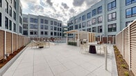 Located at 95 Anderson St in Hackensack, New Jersey, this outdoor amenity deck offers a well-appointed communal retreat set within a modern multi-story residential building. The centerpiece of the space is a generously sized, crystal-clear swimming pool, accessible via stainless steel railings and equipped with an ADA-compliant pool lift, ensuring inclusivity for all residents. Surrounding the pool is a spacious sun deck finished in large-format light stone pavers, lined with contemporary chaise lounges that invite relaxation. Warm wood-slat privacy screens define the perimeter of the deck, providing a sense of enclosure while complementing the building's sleek, contemporary façade — characterized by light gray paneling and bold-framed windows across multiple floors. A pergola-covered outdoor kitchen and grilling station serves as a social hub, complete with seating for dining and entertaining. Additional open-air lounge and dining areas are distributed across the expansive terrace, offering a variety of settings for both casual gatherings and quiet retreats. The courtyard-style layout, framed on multiple sides by the surrounding building, creates a sheltered yet open-air atmosphere that balances urban energy with resort-style comfort.