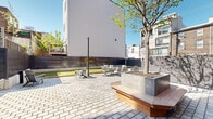 Located at 1120 Frankford Avenue in Philadelphia's dynamic Fishtown neighborhood, this thoughtfully designed outdoor amenity courtyard offers residents a well-appointed gathering space that balances recreation and relaxation. The ground-level courtyard is anchored by a full bocce ball court with a lush artificial turf surface, bordered by clean timber framing and enclosed by sleek horizontal privacy fencing — a standout feature that adds both function and a modern aesthetic. Adjacent to the bocce court, a generously sized paver patio is finished with a classic interlocking stone pattern, providing a durable and visually appealing surface throughout. The seating areas are well-furnished with Adirondack chairs, a large dining table with surrounding chairs, and additional casual seating options, making the space equally suited for socializing or quiet outdoor enjoyment. Raised concrete planters with young trees provide natural greenery and shade, softening the urban setting. String light poles are positioned throughout, suggesting an inviting ambiance during evening hours. The courtyard is framed by the building's striking mixed-material façade — combining brick and modern panel cladding — and offers views of the surrounding Fishtown streetscape, connecting residents to the neighborhood's lively character.