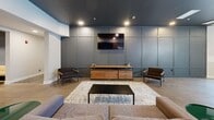 Located at 1862 E 123rd St in Cleveland, OH, this single-floor community lounge space offers a thoughtfully designed layout that balances social functionality with modern aesthetics. The open-concept kitchen is a standout feature, showcasing warm wood cabinetry, stainless steel appliances including a full-size refrigerator and wall oven, and a striking deep-blue subway tile backsplash. A large island with a crisp white countertop and bar seating serves as a central gathering point, illuminated by sleek black pendant lights that complement the polished concrete flooring. Adjacent to the kitchen, the living area features a sophisticated dark paneled accent wall with a wall-mounted flat-screen television, a wood media console, and a comfortable seating arrangement anchored by a textured area rug. The billiards area is an entertaining focal point, complete with a wood-framed pool table set beneath a dramatic ceiling installation of angled wooden slats and a cluster of globe pendant lights, adding a sculptural, artistic dimension to the space. A decorative wall-mounted display adds visual interest alongside the billiards table. Throughout, the space maintains a cohesive contemporary palette of dark and natural tones, polished concrete floors, and carefully curated lighting, creating an inviting and versatile environment well-suited for social gatherings.