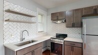 This single-floor unit offers a thoughtfully updated living space defined by classic hardwood flooring and a bright, neutral color palette throughout. The standout feature is the fully renovated kitchen, which showcases rich dark-stained shaker-style cabinetry, crisp white quartz countertops, and a striking full-wall herringbone tile backsplash that adds visual depth and character. Open floating shelves complement the cabinetry, while a stainless steel undermount sink, stainless steel electric range with hood, and a full-size refrigerator complete the functional layout. Natural light filters in through a window above the sink, enhancing the kitchen's clean, modern aesthetic. The kitchen flows openly into an adjacent living area, also finished with warm hardwood floors and natural light from a dedicated window. The bedroom features the same hardwood flooring, a built-in closet with rack storage, a window with blinds, and baseboard heating. The bathroom is finished in classic white, with small-format mosaic tile flooring accented by marble-style tile, a tiled stall shower, toilet, vanity sink, and a frosted window that brings in soft natural light while maintaining privacy. Baseboard heating is present throughout the unit.