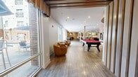Located at 1600 W Call St in Tallahassee, Florida, this thoughtfully designed single-floor amenity space offers a diverse range of social and recreational zones within a generously sized footprint. The open-concept main area features wide-plank wood-look flooring, exposed ceiling beams, and floor-to-ceiling windows along one side that invite abundant natural light and views of the surrounding exterior. A full-size pool table with a bold red felt surface anchors the game zone, complemented by an air hockey table nearby. Adjacent lounge seating — including curved caramel-toned accent chairs and a large sectional leather sofa paired with a marble-top coffee table — provides comfortable gathering spots throughout. Textured accent walls with subtle linear patterns add visual depth and a modern character to the space. A well-appointed kitchenette serves the area with warm-toned cabinetry, a beverage cooler, microwave, sink, and open shelving. A dedicated media lounge features a large-screen display, moody ceiling lighting, patterned carpet, and curated artwork with a theater theme, creating an immersive retreat. A more intimate sitting area with leather accent chairs and sophisticated décor rounds out this multifunctional community hub, making it an ideal space for socializing, recreation, and relaxation.