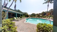 This single-story outdoor amenity facility showcases a generously sized freeform swimming pool as its centerpiece, finished in a vivid aquamarine and encircled by a broad, well-maintained brick paver deck. The pool area is equipped with a row of green lounge chairs and umbrella-shaded seating, offering ample space for relaxation under the Florida sun. A separate spa area is also present, positioned conveniently adjacent to the main pool.

The accompanying single-story clubhouse structure features a green stucco exterior with a low-pitched roof and large windows that face directly onto the pool deck, connecting the interior to the outdoor environment. A covered walkway runs along the building's poolside facade, supported by textured columns and lined with manicured shrubs, providing shaded passage between amenity areas. Spa rules and facility signage are posted near the entry doors, indicating organized management of the space.

The surrounding landscape is lush and tropical, with mature palm trees, ornamental shrubs, and flowering plants framing the pool on all sides. A pergola structure is visible at the far end of the pool area, adding an architectural focal point. The overall ambiance is bright, open, and resort-inspired, designed to maximize outdoor enjoyment in a warm climate setting.