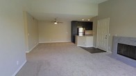 This single-floor space offers a clean, open-concept layout that seamlessly blends the living area and kitchen into one cohesive environment. The spacious living area is finished with plush wall-to-wall carpeting in a neutral tone, creating a warm and comfortable atmosphere. A ceiling fan centered overhead provides both practical air circulation and a subtle design accent.

The kitchen is defined by sleek dark cabinetry that contrasts sharply against the light-colored walls, lending a contemporary edge to the space. A raised breakfast bar with a stone-style countertop serves as a natural divider between the kitchen and living area, offering additional seating or prep space. Stainless steel appliances, including a refrigerator, complete the modern kitchen aesthetic. Recessed lighting illuminates the kitchen area, adding a polished, intentional touch to the design.

The entryway, finished with dark hardwood-style flooring, transitions smoothly into the carpeted living space, providing a clear and practical threshold. Neutral wall tones throughout the unit enhance the sense of openness and allow for versatile interior styling. The overall ambiance is modern, functional, and move-in ready, making it a well-appointed single-level living space.