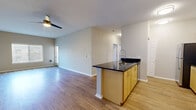 Located at 1715 E Main Ave in Puyallup, WA, this single-floor residence offers a well-designed, open-concept layout with a clean, modern aesthetic throughout. The kitchen serves as a central hub of the living space, featuring light maple cabinetry, dark granite countertops, stainless steel appliances including a refrigerator, range, and microwave, and a large peninsula island with a gooseneck faucet and undermount sink. The island provides additional seating and prep space while seamlessly connecting the kitchen to the expansive living area. The living space is finished with durable hardwood-style flooring and neutral-toned walls, creating a bright and airy atmosphere. A ceiling fan enhances comfort in the main living area, which also benefits from natural light through multiple windows. Bedrooms are carpeted for comfort and feature mirrored sliding closet doors that add depth and storage functionality. The hallway leads to a bathroom equipped with a soaking tub and shower combination set in a clean, white surround with built-in shelving. A dedicated pantry or storage closet with wire shelving offers practical organizational space. The overall layout is efficient and thoughtfully arranged, offering a comfortable and functional living environment.