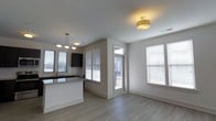 This single-floor space showcases a clean, contemporary design with an open-concept layout that connects the kitchen and living areas. Light wood-style luxury vinyl plank flooring runs throughout, providing a cohesive and modern aesthetic.

The kitchen features dark cabinetry that contrasts sharply against light countertops and a classic white subway tile backsplash. A peninsula with pendant lighting overhead defines the kitchen space while maintaining visual openness to the rest of the unit. A sink is positioned beneath a window, offering natural light while working.

The living area is well-lit by multiple windows dressed with white horizontal blinds, allowing for adjustable natural light throughout the day. A flush-mount drum light fixture with a warm gold finish serves as a subtle focal point on the ceiling. Neutral light gray walls complement the overall palette, lending the space a clean and versatile feel.

A glass-paneled exterior door with a transom window above leads to an outdoor area, further enhancing the brightness of the interior. The overall ambiance is fresh, airy, and move-in ready, reflecting a thoughtfully designed modern living environment.