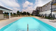 Located at 882 Taylor Station Rd in Columbus, Ohio, this outdoor amenity space centers around a generously sized swimming pool with clear, blue water. The pool area is designed for both relaxation and recreation, featuring a wide surrounding deck finished in light concrete that provides ample room for lounging and movement. Multiple chaise lounge chairs are positioned around the pool, offering comfortable spots for sunbathing or unwinding poolside. The space is enclosed by a black iron perimeter fence, providing both security and an open, unobstructed sightline to the surrounding green landscape and open skies beyond. A multi-story residential building with private balconies is situated adjacent to the pool area, and mature trees frame the perimeter, adding natural greenery to the setting. The combination of open sky, manicured grounds, and well-maintained pool infrastructure creates a resort-like atmosphere. Whether used for active swimming or leisurely relaxation, this outdoor space serves as a key communal feature of the property, offering residents a well-appointed setting to enjoy warm weather and fresh air.