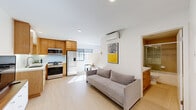 This thoughtfully designed single-floor unit offers a clean, modern aesthetic characterized by crisp white walls, light wood-toned flooring, and an open, airy layout that maximizes the sense of space throughout.

The entry hallway, equipped with a wall-mounted security keypad, leads directly into the main living area, where the kitchen, dining, and living spaces flow seamlessly together. The kitchen is well-appointed with warm wood cabinetry extending to ceiling height, a light-colored quartz countertop, a glass tile backsplash, and a full suite of stainless steel appliances including a gas range, over-the-range microwave, and a French door refrigerator with an in-door ice and water dispenser. A compact dining area with a round glass table and contemporary chair sits adjacent to the kitchen, bathed in natural light from grid-style windows.

The living area is efficiently laid out, featuring a wall-mounted split air conditioning system and space for comfortable seating. A stacked washer and dryer unit is conveniently integrated into a closeted nook within the living space.

The bathroom is finished with warm-toned ceramic tile, a soaking tub with a glass-enclosed shower, a dual-sink vanity with a white countertop, and wood cabinetry that complements the overall design palette. Recessed lighting throughout the unit adds to the clean, contemporary feel of this well-appointed space.