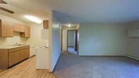 This single-floor unit offers a straightforward, open layout designed for practical living. The kitchen is positioned toward the front of the space and features white appliances, including an electric coil range, refrigerator, and dishwasher, along with wood-toned upper cabinetry and light hardwood-style flooring. A half-wall partition separates the kitchen from the adjacent living area, which transitions to wall-to-wall carpet that extends throughout the main living space and down the hallway.

The living area is modest in scale with neutral light blue-grey walls and a flush-mount ceiling light that provides general illumination. A hallway leads further into the unit, where at least one additional room is visible through a dark wood-trimmed doorway, suggesting a bedroom or additional living space. The corridor layout provides a clear sense of flow through the unit.

Overall, the space presents a clean, utilitarian design with consistent neutral tones throughout. The combination of hard flooring in the kitchen and carpet in the living and hallway areas offers a functional transition between zones suited for everyday use.
