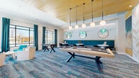 Located at 8707 N Davis Hwy in Pensacola, Florida, this well-appointed community game and lounge room offers a thoughtfully designed recreational space ideal for socializing and entertainment. The single-floor layout makes efficient use of its generously sized footprint, creating a comfortable and inviting atmosphere throughout.

The space is anchored by a contemporary light wood pool table positioned beneath a striking row of globe pendant lights suspended from a warm wood-paneled ceiling. A foosball table adds to the entertainment options, while a large wall-mounted flat-screen TV is set against a decorative herringbone-patterned accent wall, adding visual texture to the room.

A teal-upholstered banquette lines one wall, complemented by round white café tables and modern black swivel chairs — an ideal setting for casual gatherings. Decorative circular mirrors and teal chandelier accents above this seating zone add a playful, artistic flair. Opposite, a lounge seating area features white armchairs with teal cushions arranged around a geometric side table, bathed in natural light from oversized black-framed windows draped with rich teal curtains.

Coastal-inspired artwork, including a large ocean wave canvas, reinforces the breezy, resort-like ambiance. The multi-toned blue and gray patterned carpet ties the entire space together, evoking the feel of the nearby Gulf Coast.
