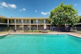 Located at 915 S College Rd in Lafayette, Louisiana, this outdoor pool area serves as a central amenity within a two-story brick and stucco building complex. The courtyard-style layout positions a generously sized rectangular swimming pool as the focal point, its clear turquoise water reflecting the Louisiana sunshine. The pool is accessible via built-in entry steps and is encircled by a spacious concrete and paver deck, providing ample room for relaxation and recreation.

The surrounding deck is furnished with lounge chairs, canopied sun beds, and rattan seating arrangements, offering multiple areas to unwind in the open air. A wrought iron perimeter fence encloses the pool area, adding a layer of security while maintaining an open, airy feel. Mature trees at the corners of the courtyard provide natural shade and a touch of greenery against the warm Louisiana sky.

The building that frames the courtyard features two stories of covered walkways and private balconies accessible from individual units, creating an inward-facing design that overlooks the pool. The classic brick construction at ground level, combined with white-painted columns and railings on the upper floor, reflects a traditional Southern architectural style. The entire space is bathed in natural light, making it an inviting and practical outdoor gathering area.