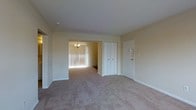 This single-floor space presents a clean, functional layout with a neutral color palette of soft beige tones on both the walls and ceiling, creating a calm and cohesive atmosphere throughout. The main living area is finished with wall-to-wall carpet in a light tan shade, lending the space a comfortable, understated feel.

A set of bi-fold closet doors provides convenient built-in storage, positioned adjacent to the unit's entry door, which features a multi-lock setup for added security. The front entry area transitions naturally into the main living space without obstruction, contributing to an open and accessible flow.

An open doorway connects the living area to an adjoining room, visible in the background and illuminated by a classic chandelier. That secondary room benefits from substantial natural light filtering in through floor-length vertical blinds, brightening the overall interior. The carpeting continues seamlessly between both rooms, reinforcing the sense of a unified, flowing layout. White baseboards frame each room, adding a crisp finishing detail against the warm wall tones. Overall, the space offers a straightforward, move-in-ready environment with practical storage and good connectivity between rooms.