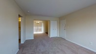 This single-floor space presents a clean, functional layout with a neutral color palette of soft beige tones on both the walls and ceiling, creating a calm and cohesive atmosphere throughout. The main living area is finished with wall-to-wall carpet in a light tan shade, lending the space a comfortable, understated feel.

A set of bi-fold closet doors provides convenient built-in storage, positioned adjacent to the unit's entry door, which features a multi-lock setup for added security. The front entry area transitions naturally into the main living space without obstruction, contributing to an open and accessible flow.

An open doorway connects the living area to an adjoining room, visible in the background and illuminated by a classic chandelier. That secondary room benefits from substantial natural light filtering in through floor-length vertical blinds, brightening the overall interior. The carpeting continues seamlessly between both rooms, reinforcing the sense of a unified, flowing layout. White baseboards frame each room, adding a crisp finishing detail against the warm wall tones. Overall, the space offers a straightforward, move-in-ready environment with practical storage and good connectivity between rooms.