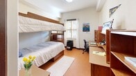 This 130 square foot dorm room, located at 2299 Piedmont Ave in Berkeley, CA, exemplifies efficient student living. The space is thoughtfully designed with built-in wooden furniture that maximizes the compact area. A bunk bed dominates one wall, providing sleeping quarters for two occupants. Adjacent to the beds, a well-organized study area features a long desk with multiple drawers and shelving units above, offering ample storage for books and personal items. The room boasts a large window with blinds, allowing natural light to brighten the space and providing ventilation. Cork flooring adds warmth and durability to the room. The entrance features a solid wood door with modern security features. Wall decor, including a UC Berkeley pennant and a Bay Area poster, adds a personalized touch to the academic environment. Despite its small size, the room's layout creates distinct zones for sleeping, studying, and storage, making it a functional living space for university students.
