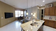This single-story residence showcases an open-concept layout that seamlessly connects the kitchen and living areas, creating a cohesive and functional living environment. The kitchen features a prominent granite-topped island with a double stainless steel sink, complemented by dark cabinetry below. Three elegant pendant lights hang above the island, providing focused task lighting while adding a decorative element to the space.

The kitchen flows naturally into the living area, which is anchored by a neutral-toned sofa, a glass coffee table, and a patterned area rug. A wall-mounted television and a console table with decorative accessories complete the living space. Large windows fitted with horizontal blinds invite natural light, brightening the room and enhancing the airy atmosphere.

Warm taupe walls and large-format polished tile flooring run throughout the space, providing a cohesive aesthetic. Recessed ceiling lighting supplements the natural light, ensuring consistent illumination throughout. A doorway visible in the background hints at additional rooms beyond. The overall design balances contemporary style with a warm, welcoming ambiance.