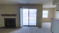 This single-floor residential space offers a comfortable and functional layout designed for everyday living. The main living area is carpeted throughout, providing a warm and cozy atmosphere underfoot. A tile-surround fireplace with a classic wood mantel serves as a natural focal point, adding both character and practicality to the room.

Natural light flows generously into the space through a large sliding glass door, which opens directly to a patio area and offers views of the surrounding neighborhood. An additional window on the adjacent wall further brightens the room, enhancing the open and airy feel of the interior. Sheer panel curtains frame the sliding door, offering a light touch of softness to the space.

The walls are finished in a neutral beige tone, complementing the light-colored carpet and creating a versatile backdrop suitable for a variety of interior styles. A textured ceiling and visible HVAC vent overhead reflect a practical, well-maintained construction. The overall ambiance is understated and move-in ready, offering a clean, functional environment with key amenities and easy indoor-outdoor access through the sliding patio door.