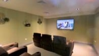 This single-floor space is purpose-built for entertainment, centered around a private home theater room that delivers a true cinematic atmosphere. The room is finished with smooth, light-colored walls and recessed ceiling details that lend a clean, polished aesthetic throughout. A large wall-mounted flat-screen display serves as the focal point, positioned for optimal sightlines from every seat in the room. The seating arrangement features multiple rows of plush, dark-upholstered recliner chairs, providing generous comfort for an audience of family and guests alike. The tiered layout ensures unobstructed views of the screen from all positions, mimicking the feel of a professional movie theater. Ceiling-mounted speakers suggest an integrated audio setup designed to complement the visual experience with immersive sound. Subtle ambient lighting along the lower walls adds a tasteful, theater-inspired glow without detracting from the on-screen presentation. The overall ambiance is intimate and refined, making this space an ideal retreat for movie nights, sports viewing, or gaming. Every design element has been thoughtfully chosen to prioritize comfort, functionality, and a high-quality entertainment experience.