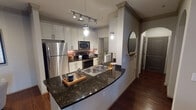 Located at 5201 Collin-McKinney Pkwy in McKinney, TX, this single-floor residence combines modern finishes with thoughtfully designed living spaces. The kitchen serves as a functional centerpiece, showcasing dark granite countertops with a curved peninsula, white shaker cabinetry, stainless steel appliances including a full-size refrigerator and built-in microwave, a subway tile backsplash, and a dedicated laundry area. Track lighting and pendant fixtures provide targeted illumination throughout. The living room impresses with a tray ceiling, rich hardwood flooring, a bold teal accent wall, and access to an outdoor balcony through full-length glass doors that invite natural light. Each bedroom features plush carpeting, large windows with blinds, and distinct accent walls — one in deep navy and another in rich emerald green — creating striking, individual character. Walk-in closets provide ample storage. The bathrooms are finished with granite vanity tops, tile flooring, and tub-shower combinations with subway tile surrounds and large mirrors. The cohesive design throughout the residence balances bold color choices with refined architectural details, resulting in a dynamic and comfortable living environment.