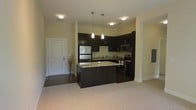 This single-floor residence offers a thoughtfully designed open-concept layout that seamlessly connects the kitchen and living areas. The kitchen serves as the focal point of the space, showcasing rich dark espresso cabinetry that provides ample storage and a bold contrast against the light-toned walls and flooring. A central kitchen island with granite countertops offers both additional prep space and a natural gathering point. Stainless steel appliances, including a refrigerator, microwave, and range, lend a sleek, contemporary feel to the cooking space. Elegant pendant lights hang above the island, complementing the flush-mount ceiling fixtures that provide consistent, well-distributed illumination throughout. The transition from the hard flooring near the entryway to the plush carpeting in the living area creates a clear yet fluid delineation between functional zones. The entryway is clean and welcoming, with a classic white door and neutral walls. Overall, the space maintains a modern aesthetic with a warm, neutral color palette that balances practicality with inviting comfort, making it well-suited for everyday living and casual entertaining alike.