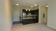 This single-floor residence offers a thoughtfully designed open-concept layout that seamlessly connects the kitchen and living areas. The kitchen serves as the focal point of the space, showcasing rich dark espresso cabinetry that provides ample storage and a bold contrast against the light-toned walls and flooring. A central kitchen island with granite countertops offers both additional prep space and a natural gathering point. Stainless steel appliances, including a refrigerator, microwave, and range, lend a sleek, contemporary feel to the cooking space. Elegant pendant lights hang above the island, complementing the flush-mount ceiling fixtures that provide consistent, well-distributed illumination throughout. The transition from the hard flooring near the entryway to the plush carpeting in the living area creates a clear yet fluid delineation between functional zones. The entryway is clean and welcoming, with a classic white door and neutral walls. Overall, the space maintains a modern aesthetic with a warm, neutral color palette that balances practicality with inviting comfort, making it well-suited for everyday living and casual entertaining alike.