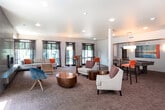 This well-appointed single-floor clubhouse offers an expansive, open-concept layout designed for comfort and social interaction. The main lounge area is generously sized, featuring plush seating arrangements with neutral-toned sofas and armchairs accented by warm orange throw pillows. A floor-to-ceiling tiled fireplace serves as a striking focal point, flanked by large windows dressed in dark curtain panels that frame views of the surrounding greenery, filling the space with abundant natural light.

The lounge transitions seamlessly into a casual gathering zone with high-top bar tables and stools, ideal for informal meetings or socializing. A billiards table is also present, adding a recreational dimension to the space. Recessed lighting and arc floor lamps provide warm, layered illumination throughout.

The full kitchen is a standout feature, boasting a large central island with a smooth countertop, stainless steel appliances including a French door refrigerator and double wall oven, contemporary cabinetry in light and dark wood tones, a tile backsplash, and pendant lighting overhead. A wall of sliding glass doors connects the kitchen to an outdoor deck area, enhancing the flow between interior and exterior.

The entire space is finished with patterned carpeting and neutral wall tones, creating a cohesive, modern, and inviting atmosphere throughout.