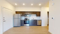 This single-floor unit offers a clean, modern design with light hardwood-style flooring and neutral tones throughout. The kitchen is a standout feature, equipped with a full suite of stainless steel appliances including a refrigerator, electric range, over-the-range microwave, and dishwasher. Dark espresso-toned cabinetry lines the walls from floor to ceiling, complemented by granite-style countertops and a classic white subway tile backsplash. Recessed lighting illuminates the open layout, which connects the kitchen seamlessly to the living area. The bathroom is both functional and well-designed, featuring a dark wood vanity with a granite-style countertop, a large mirror, and a full soaking tub with shower. Natural light enters through windows fitted with blinds, and a stacked washer and dryer unit is conveniently integrated into the bathroom, offering in-unit laundry. The bedroom is spacious and bright, with a large window providing views of the surrounding greenery and ample wall space for flexible furnishing. Throughout the unit, consistent light wood flooring and white walls create a bright, cohesive atmosphere that feels open and welcoming.